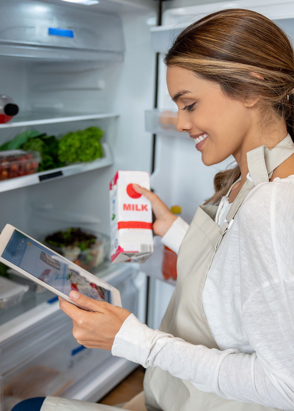 Woman holding a milk carton in front of an open refrigerator while reading a recipe on a tablet (who does that? seriously, stock photos are weird)